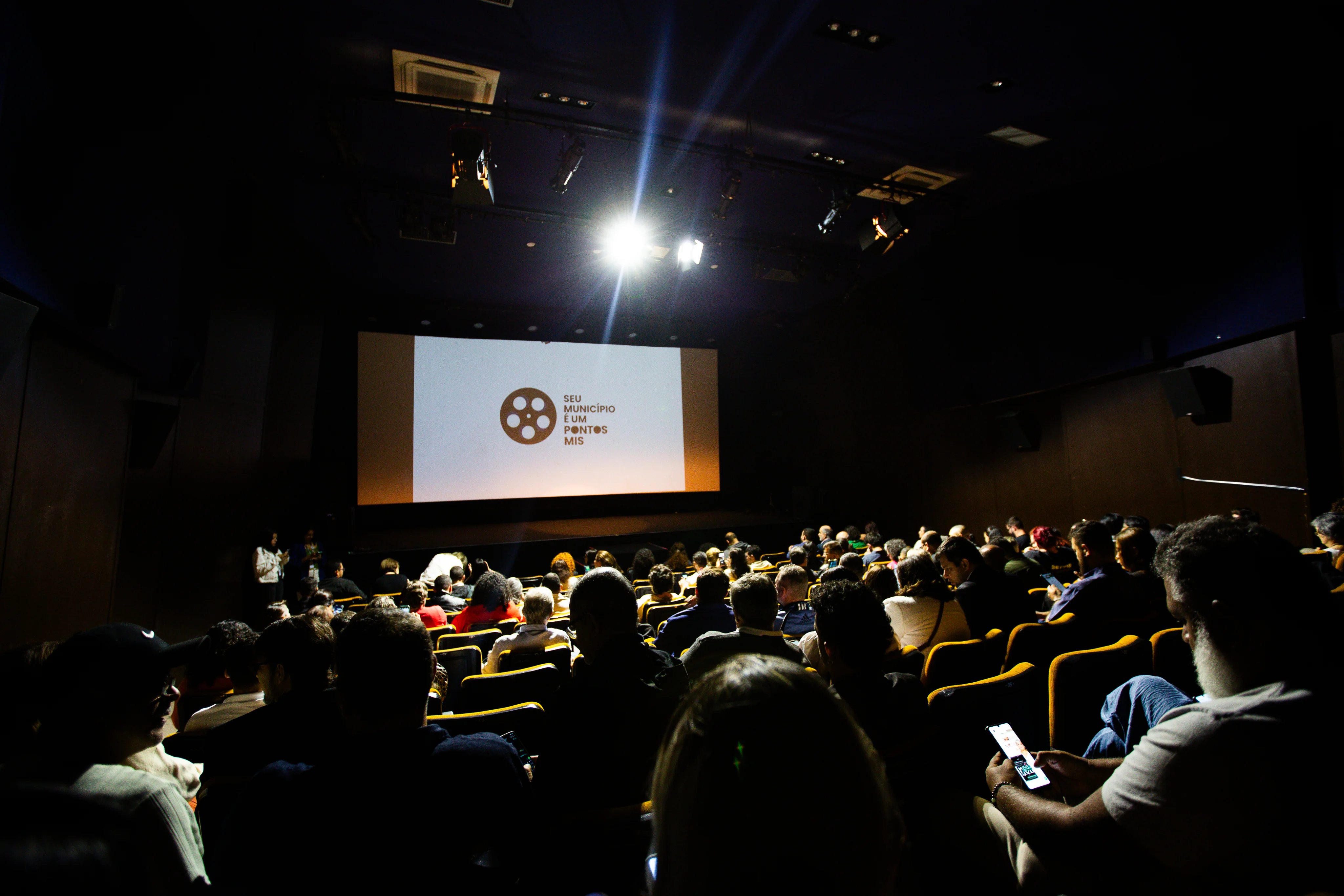 Uma sala de cinema com várias pessoas assistindo, e na tela, escrito 'Seu municipio é um ponto MIS'